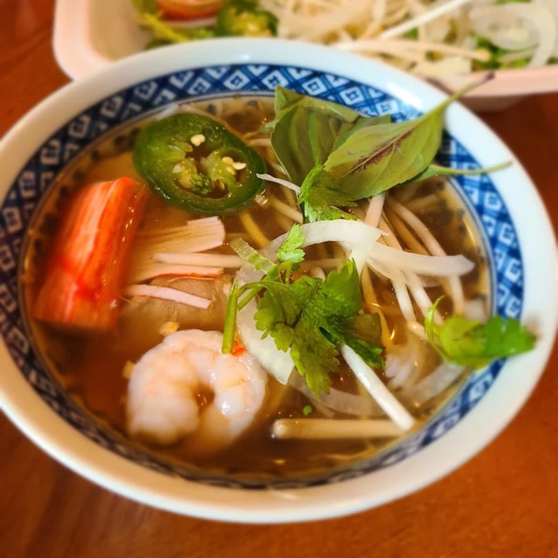 Close-up of a Thai noodle soup bowl with shrimp, crab stick, jalapenos, and herbs.
