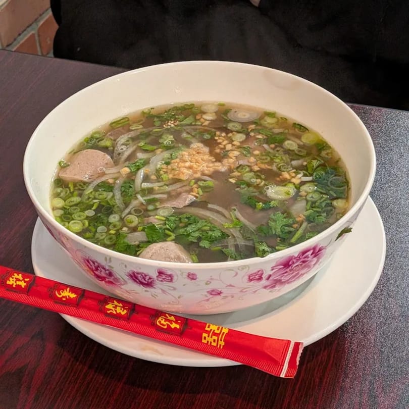 A bowl of Thai pho soup with meat, green onions, and cilantro, with red chopsticks beside it.
