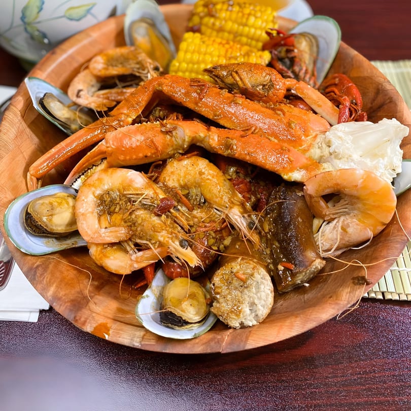 A large wooden bowl filled with a Thai seafood boil including crab legs, shrimp, mussels, and corn.